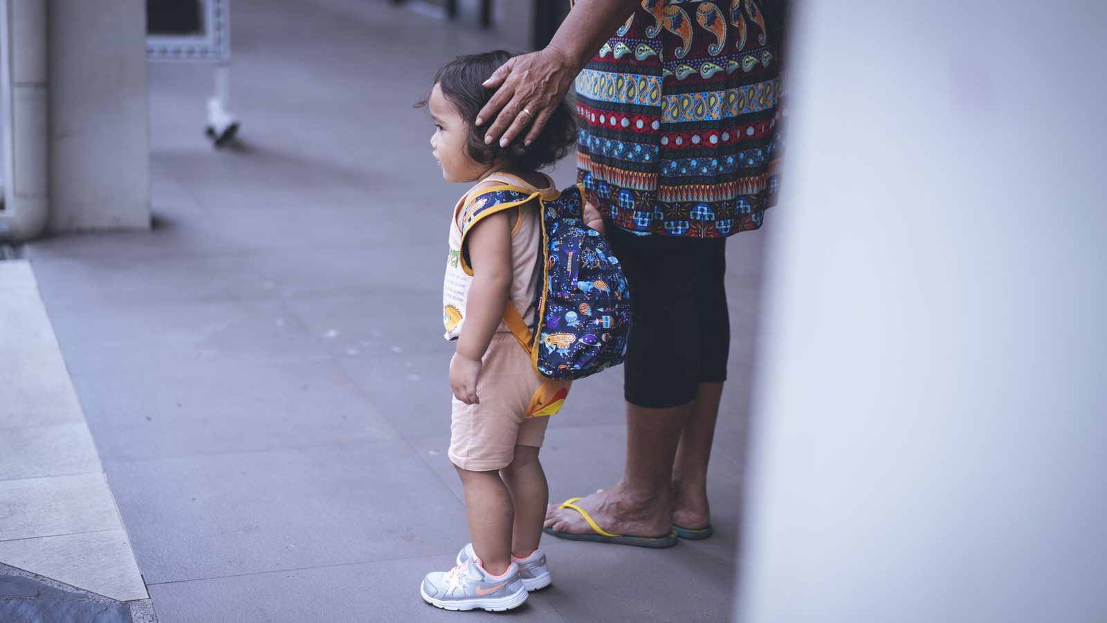 Criança pequena de mochila nas costas ao lado de um adulto, que apoia a mão na cabeça dela